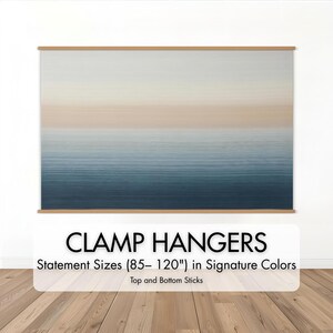 May include: A large, abstract artwork with a gradient of colors from light beige to deep blue, displayed with wooden clamp hangers. The text "CLAMP HANGERS" and "Statement Sizes (85-120") in Signature Colors" is visible.