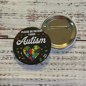 May include: A black button with a white heart made of puzzle pieces and the text "Please be patient I have Autism". The button has a silver pin back.