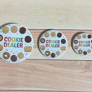 May include: Three round white buttons with a variety of cookies around the edge. The text "COOKIE DEALER" is in rainbow colors in the center. The buttons are on a light brown wooden surface.