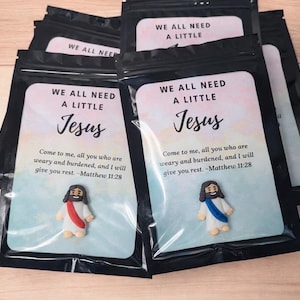 May include: Several black pouches with a printed card featuring the text "WE ALL NEED A LITTLE Jesus". The card includes a quote from Matthew 11:28 and a small figurine of Jesus. The background is a light blue and pink watercolor design.