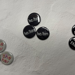 May include: A collection of pronoun buttons. Three black buttons with "he/him" in white, three with "they/them", and three gray buttons with "she/her". The buttons are arranged on a textured, off-white surface.