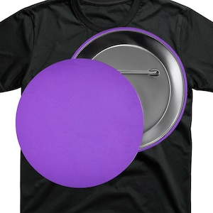 May include: A black t-shirt with a large, round, purple button pinned to it. The button is a solid, vibrant purple color and has a silver backing with a pin. The button is partially covering the shirt.