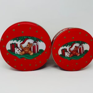 May include: Two round red Christmas tins with a green holly pattern and a bear wearing a Santa hat and holding presents.