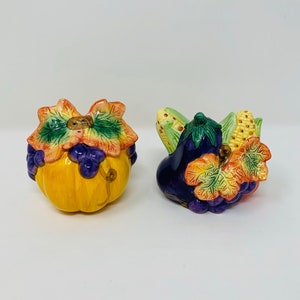 May include: A set of two ceramic salt and pepper shakers shaped like a pumpkin and an eggplant. The pumpkin shaker is yellow with green leaves and purple grapes. The eggplant shaker is purple with green leaves and yellow corn.