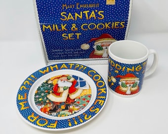 May include: A Mary Engelbreit Santa's Milk & Cookies Set, featuring a blue box, a plate, and a mug. The plate and mug have a festive design with Santa Claus. The plate reads "ME??!! COOKIES FOR SANTA!!!".