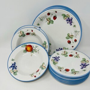 May include: A set of white ceramic dinnerware with a blue rim and a fruit pattern featuring strawberries, grapes, and apples. The set includes a large dinner plate, a smaller salad plate, and a soup bowl.