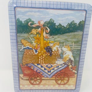 May include: A rectangular tin box with a painted scene of a picnic basket on a red wagon, with two cats. The basket is filled with food and flowers. The box has a blue border with a striped pattern.