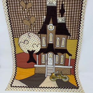 May include: A brown and beige quilted wall hanging featuring a brown house with white windows and a white door. The house has a brown roof with white trim and a white chimney. There is a brown tree with white flowers and two brown butterflies in the background. The house is on a yellow background with brown and white polka dots.