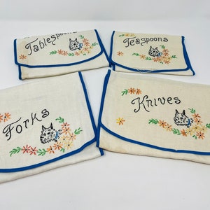 May include: Four white fabric pouches with blue trim and embroidered floral designs. Each pouch has a different category of silverware embroidered on it: "Tablespoons", "Teaspoons", "Knives", and "Forks".