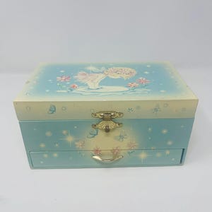May include: A rectangular light blue jewelry box with a hinged lid and a single pull-out drawer. The exterior features a painted illustration of a small figure with wings, flowers, and butterflies, accented with gold-toned metal hardware.