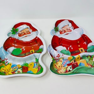 May include: Two white ceramic Santa Claus serving dishes with red and green accents. Each dish features a Santa Claus holding a candy cane and a bag full of presents, including a teddy bear, a toy duck, and a toy train.