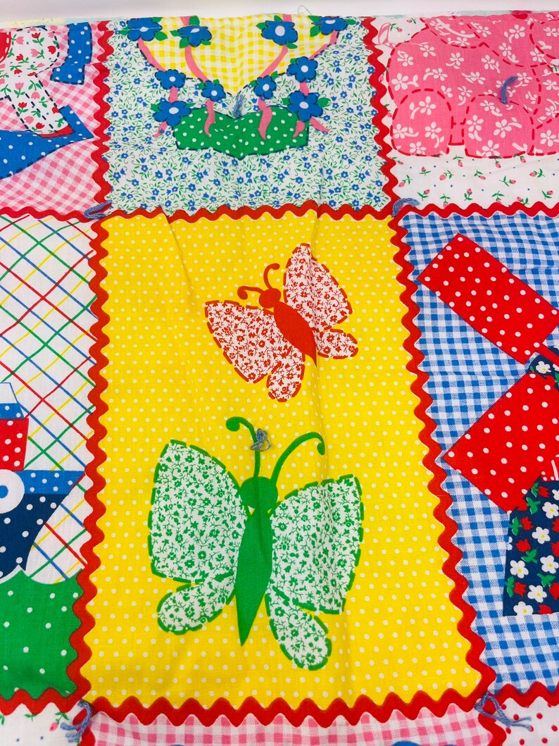 May include: A yellow fabric with a red, white, and blue checkered pattern. The fabric has a scalloped edge and is decorated with two butterflies, one green and one red. The butterflies are made of fabric with a floral pattern.