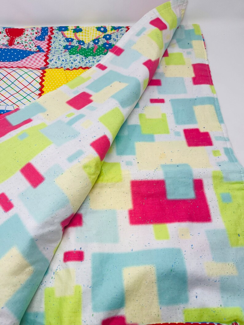 May include: A white blanket with a geometric pattern of pink, blue, and green squares. The blanket is folded in half and the pattern is visible on both sides.