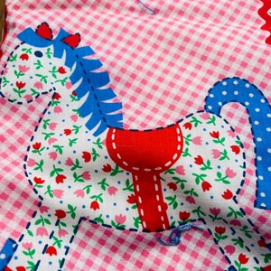 May include: A patchwork fabric rocking horse with a blue mane, red saddle, and white floral print. The horse is on a pink and white gingham background.