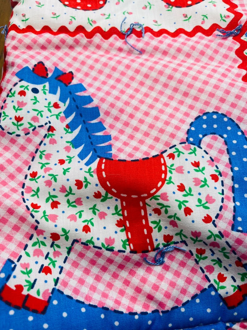 May include: A patchwork fabric rocking horse with a blue mane, red saddle, and white floral print. The horse is on a pink and white gingham background.