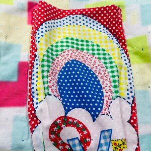 May include: A white fabric patch with a rainbow design in red, yellow, green, blue, and white. The rainbow is surrounded by a white cloud with a red, white, and blue flower design. The flower has a red center with white flowers and a blue and white checkered "V" in the center.