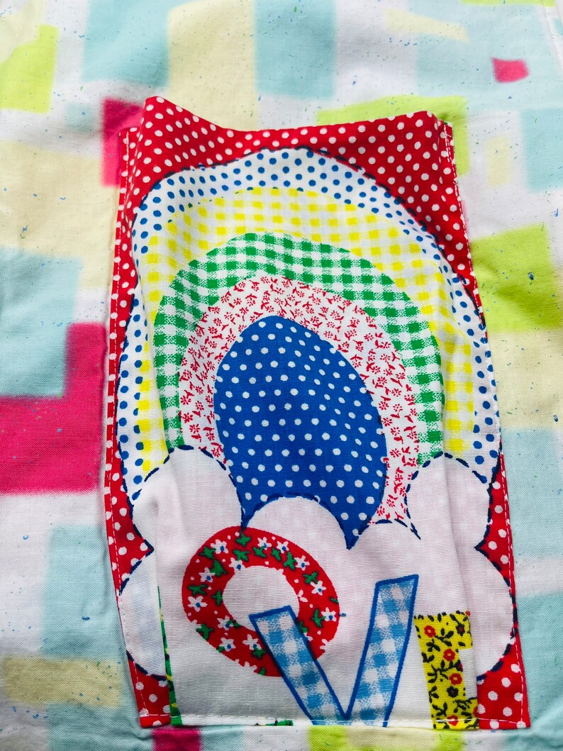 May include: A white fabric patch with a rainbow design in red, yellow, green, blue, and white. The rainbow is surrounded by a white cloud with a red, white, and blue flower design. The flower has a red center with white flowers and a blue and white checkered "V" in the center.