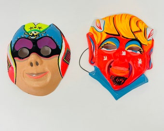 May include: Two vintage Halloween masks. One is a blue, yellow, and purple superhero mask with a skull and crossbones design. The other is a red, yellow, and blue devil mask with horns.