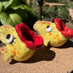 May include: Two ceramic fish figurines with open red mouths, yellow bodies, and orange spots. The fish have large, black and white eyes and yellow fins. They are displayed on a brick surface with green foliage in the background.