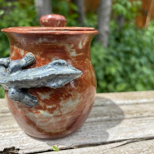 May include: A brown ceramic jar with a lid and a gray ceramic lizard design. The jar has a speckled glaze and the lizard is partially wrapped around the jar.