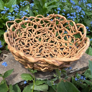 Peut inclure: Un bol en céramique brun avec un motif tissé, ressemblant à un nid ou à un panier. Le bol est posé sur un rocher avec des fleurs bleues en arrière-plan.