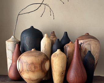 May include: A collection of various wooden vases in different shapes, sizes, and wood tones, including light tan, reddish-brown, and matte black finishes. One tall, light-colored vase holds a single, thin, curved, leafless branch.