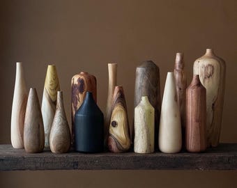 May include: A collection of wooden vases in various shapes, sizes, and wood grains. The vases are arranged on a wooden shelf. Some of the vases are natural wood color, while others are stained in shades of brown, black, and green.