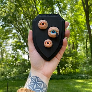 May include: A black wooden plaque with three decorative eyes. The eyes are made of clay and have different colors, including blue, orange, and brown.