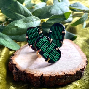 May include: Three black rings with gold accents. The rings are oval shaped and feature a green binary code pattern. The rings are displayed on a wooden surface.