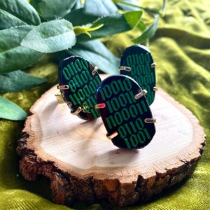 Op de afbeelding: Drie zwarte ovale ringen met gouden accenten. De ringen zijn gemaakt van steen en hebben een groen binair codepatroon.