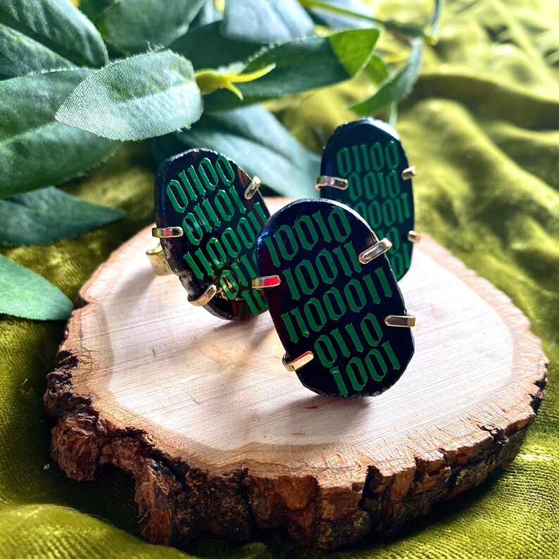 Op de afbeelding: Drie zwarte ovale ringen met gouden accenten. De ringen zijn gemaakt van steen en hebben een groen binair codepatroon.