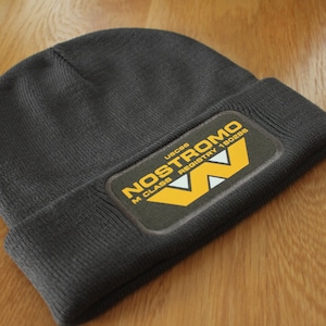 May include: A dark grey beanie with a black and yellow patch that reads "Nostromo M Class Registry 180888" and "USCSS".