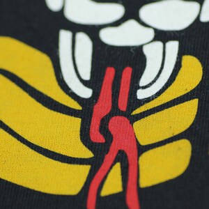 May include: Close-up of a black fabric with a yellow, red, and white graphic design. The design features a stylized figure with a long, red tongue.