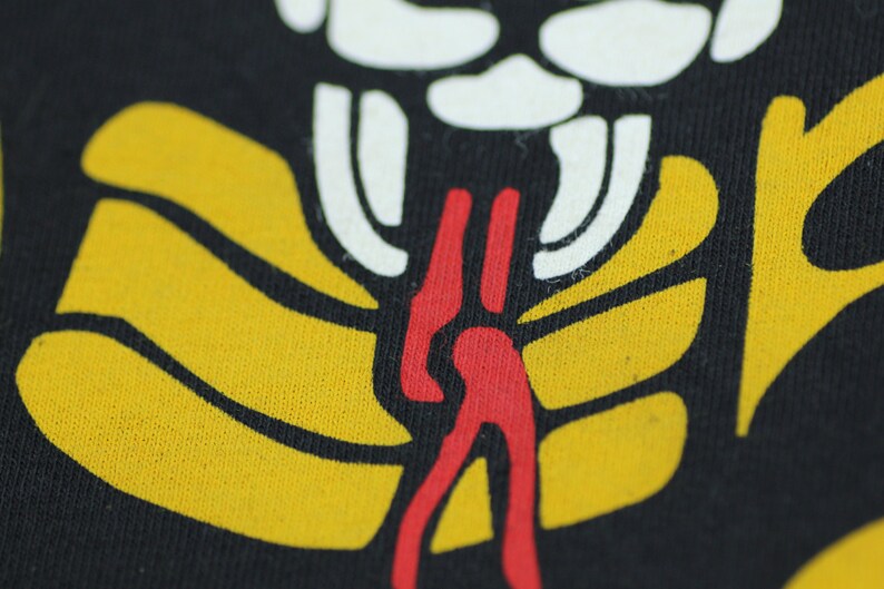 May include: Close-up of a black fabric with a yellow, red, and white graphic design. The design features a stylized figure with a long, red tongue.