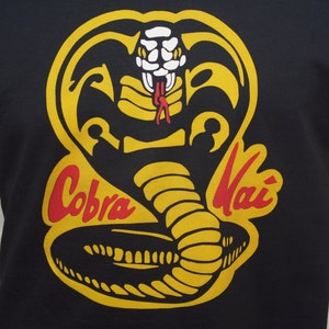 May include: Black t-shirt with a yellow and red Cobra Kai logo. The logo features a cobra with its head raised and fangs showing. The words "Cobra Kai" are written in red and yellow.