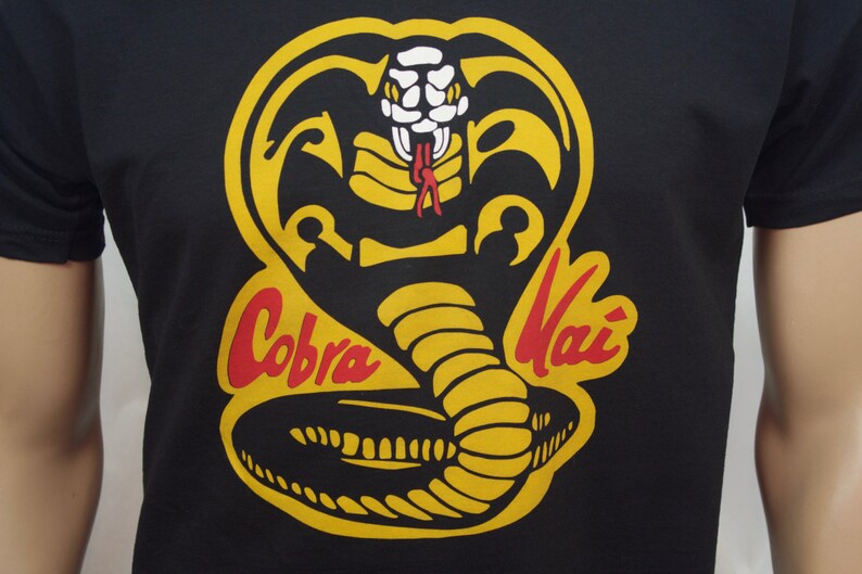 May include: Black t-shirt with a yellow and red Cobra Kai logo. The logo features a cobra with its head raised and fangs showing. The words "Cobra Kai" are written in red and yellow.