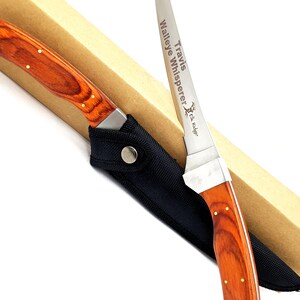 Puede incluir: Un cuchillo de caza grabado personalizado con mango de madera y funda negra. La hoja est&aacute; grabada con el texto "Travis' Walleye Whisperer Elk Ridge".