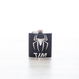 K&ouml;nnte beinhalten: Schwarze Flasche mit einem silbernen Spinnenmotiv und dem Namen "TIM" auf der Vorderseite.