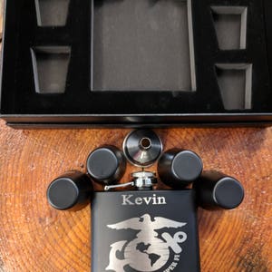May include: Black flask with a silver lid and engraved text "Kevin Best Man 3-18-18". The flask features a silver eagle and globe with the words "Semper Fi" underneath. The flask is surrounded by four black shot glasses and a silver funnel.