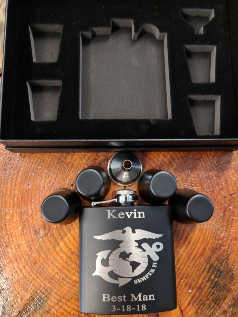 May include: Black flask with a silver lid and engraved text "Kevin Best Man 3-18-18". The flask features a silver eagle and globe with the words "Semper Fi" underneath. The flask is surrounded by four black shot glasses and a silver funnel.