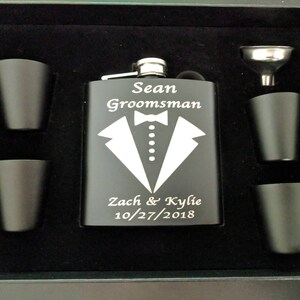 May include: Black flask with a silver top and engraved text: "Sean Groomman Zach & Kylie 10/27/2018". The flask is surrounded by four small black shot glasses and a silver funnel.