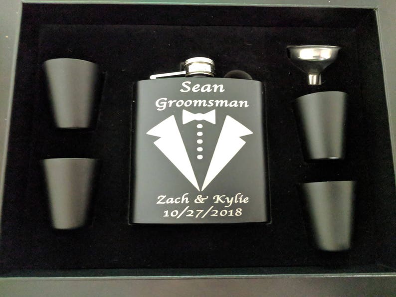 May include: Black flask with a silver top and engraved text: "Sean Groomman Zach & Kylie 10/27/2018". The flask is surrounded by four small black shot glasses and a silver funnel.