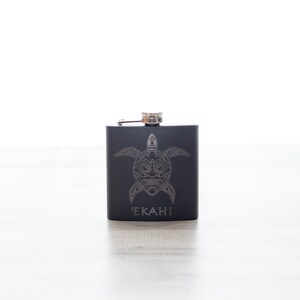 K&ouml;nnte beinhalten: Schwarze Metallflasche mit einem silbernen Schildkr&ouml;ten-Design und dem Wort "EKAHI" auf der Vorderseite.