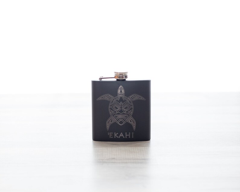 K&ouml;nnte beinhalten: Schwarze Metallflasche mit einem silbernen Schildkr&ouml;ten-Design und dem Wort "EKAHI" auf der Vorderseite.