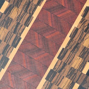 May include: A wooden chopping board with a chevron pattern in red and brown wood. The board is also made with a light wood and a dark wood with a black grain.