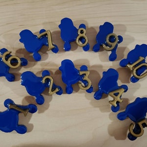 May include: A set of 10 blue poodle-shaped dice with gold numbers 1 through 10. The dice are arranged in a semi-circle on a light wood surface.
