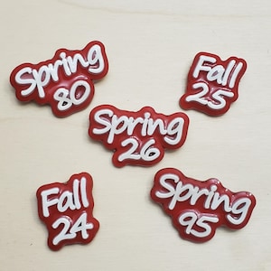 May include: Five red enamel pins with white raised lettering. The pins feature the text Spring 80, Fall 25, Spring 26, Fall 24, and Spring 95. Each pin has a glossy finish and irregular, rounded edges that follow the shape of the white text.