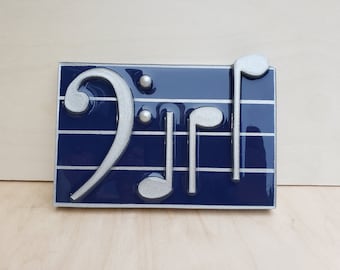 May include: A rectangular plaque featuring musical notes and symbols. The design includes a silver treble clef, quarter notes, and a bass clef, set against a dark blue background with silver lines. The plaque is likely a decorative item.