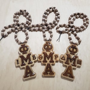 Puede incluir: Tres collares de madera con un colgante de cruz que presenta las letras "ΦΜΑ" en un esquema de color marrón y dorado. Los collares están hechos de cuentas de madera marrón.