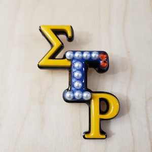 May include: A gold and black sorority pin with the letters "ΣΓΡ" in yellow and black. The "Γ" is decorated with white pearls.